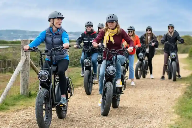 A group of seven people riding electric bikes on a gravel path, smiling and chatting. They are wearing helmets and casual clothing, with scenic hills in the background.