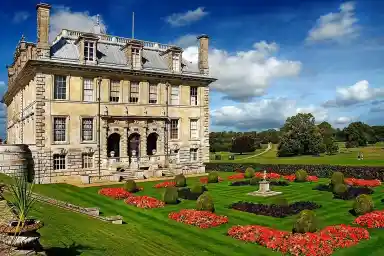 A grand, historic mansion with a slate roof and ornate facade is surrounded by neatly manicured gardens featuring colorful flower beds and sculpted hedges. Lush greenery and a wide pathway extend into the distance under a partly cloudy sky.