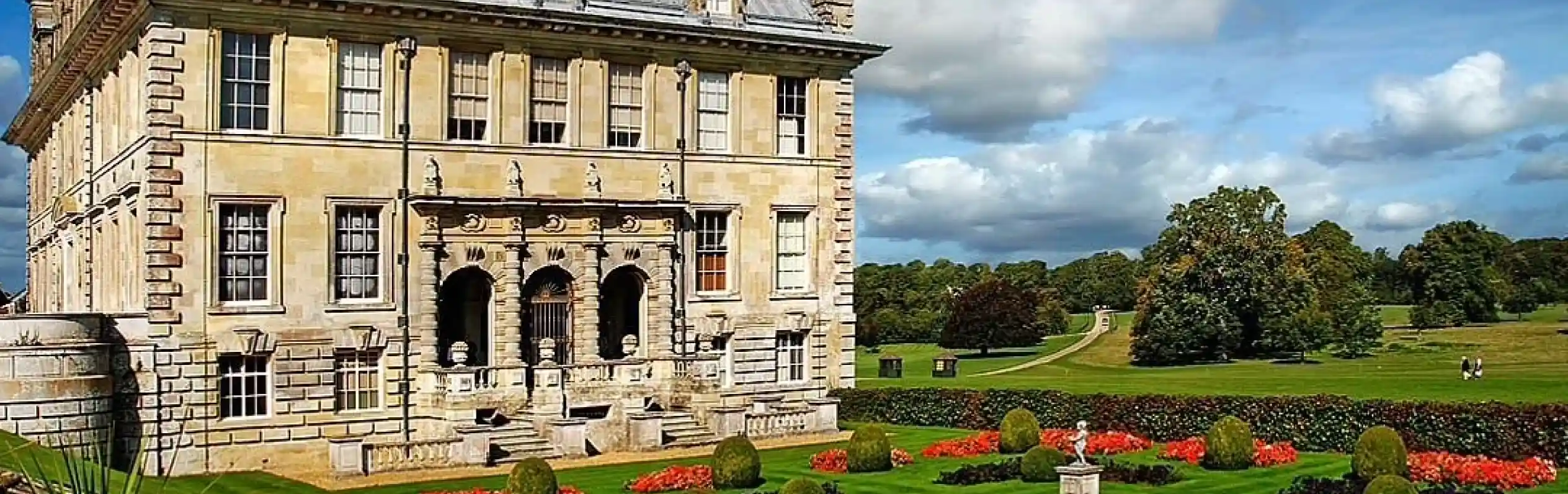 A grand, historic mansion with a slate roof and ornate facade is surrounded by neatly manicured gardens featuring colorful flower beds and sculpted hedges. Lush greenery and a wide pathway extend into the distance under a partly cloudy sky.
