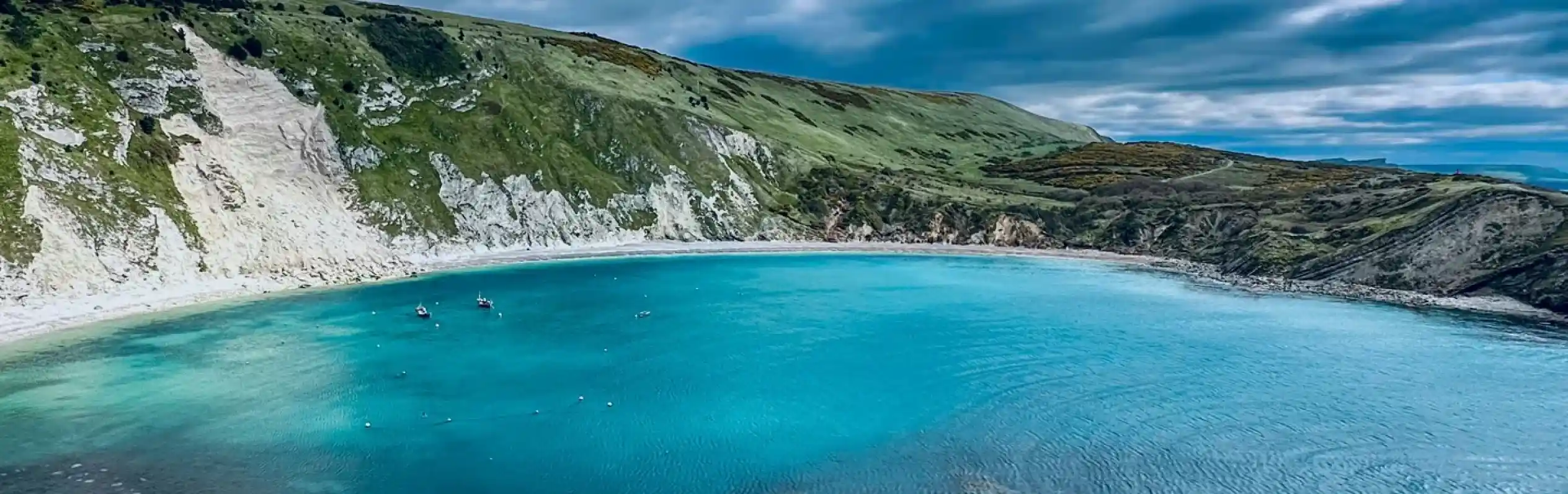 A serene coastal landscape featuring a clear turquoise bay surrounded by steep, grassy cliffs under a cloudy sky. Several small boats are anchored in the calm waters.