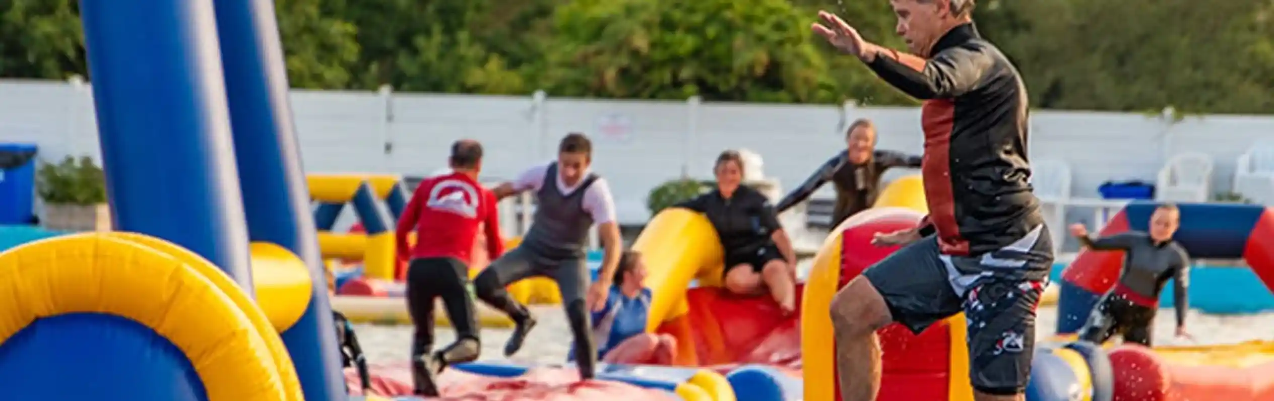 A man in a black shirt and shorts balances on a large inflatable structure at a water park. Others can be seen navigating various colorful inflatable obstacles in the background. Trees and a clear sky are visible, indicating a sunny day.