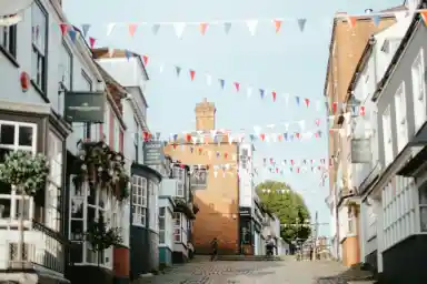 A quaint street lined with charming buildings, adorned with red, white, and blue bunting. Cobblestone path leads to a slightly elevated area with a tower in the distance.