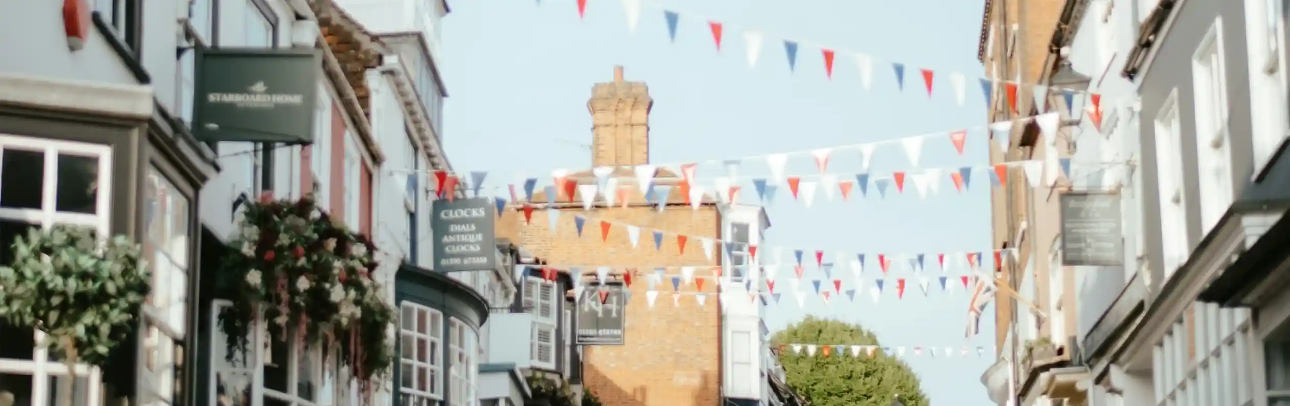 A quaint street lined with charming buildings, adorned with red, white, and blue bunting. Cobblestone path leads to a slightly elevated area with a tower in the distance.