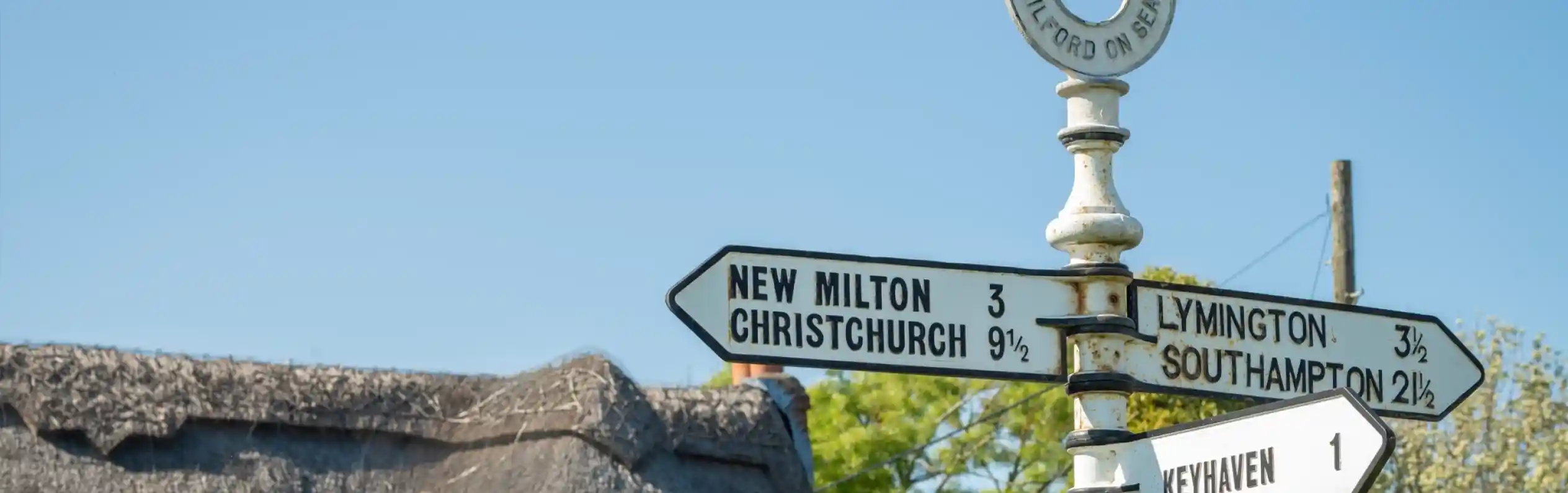 A vintage-style direction signpost with multiple arrows indicating distances to various locations: New Milton (3 miles), Christchurch (9.1 miles), Lymington (3.5 miles), Southampton (2.5 miles), and Keyhaven (1 mile). A thatched roof structure is visible in the background against a clear blue sky.