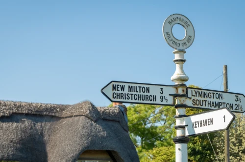 A vintage-style direction signpost with multiple arrows indicating distances to various locations: New Milton (3 miles), Christchurch (9.1 miles), Lymington (3.5 miles), Southampton (2.5 miles), and Keyhaven (1 mile). A thatched roof structure is visible in the background against a clear blue sky.