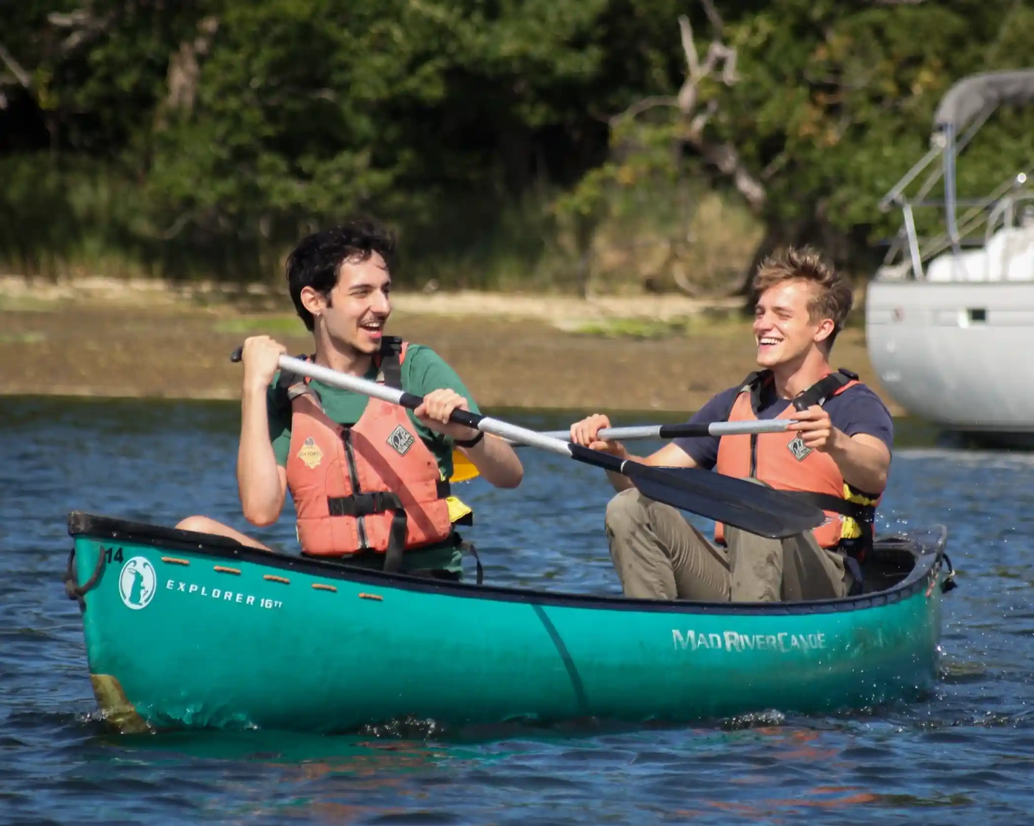 Two young men paddling together in a green canoe on a calm body of water. Both are wearing orange life jackets and smiling at each other, enjoying a sunny day outdoors. In the background, trees and a boat are visible.