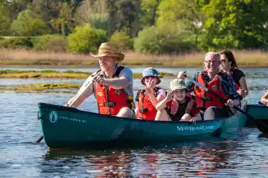 A family enjoys a sunny day canoeing on a calm river. Two adults and two children wear life jackets and are smiling while paddling. Lush greenery and reeds line the riverbank in the background.