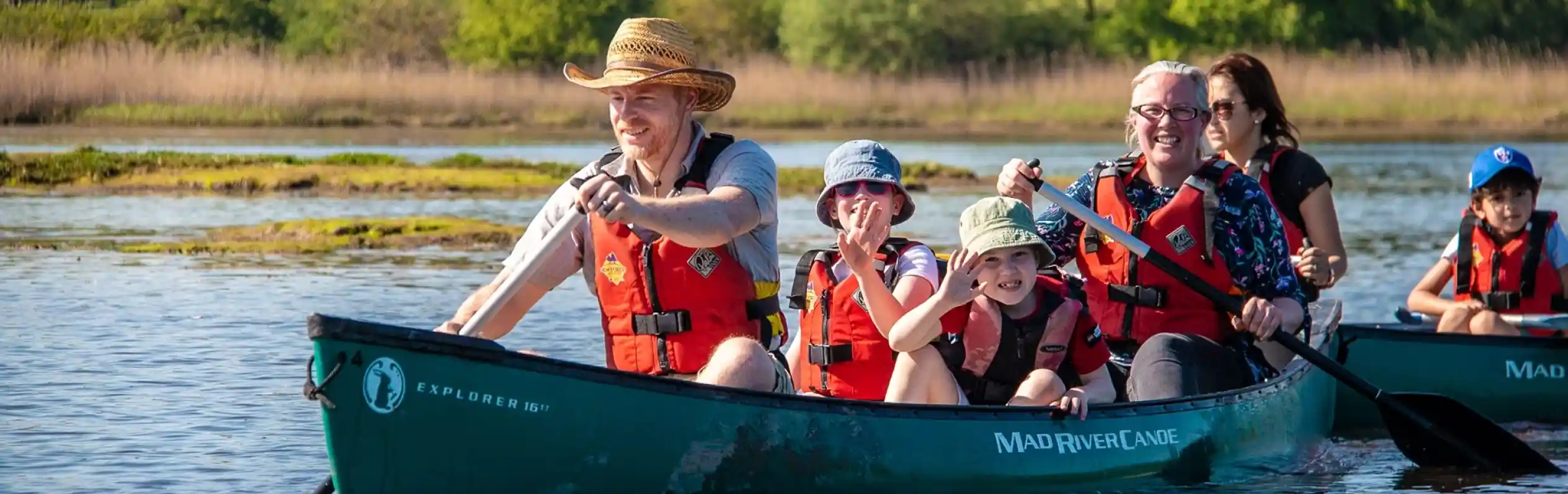 A family enjoys a sunny day canoeing on a calm river. Two adults and two children wear life jackets and are smiling while paddling. Lush greenery and reeds line the riverbank in the background.