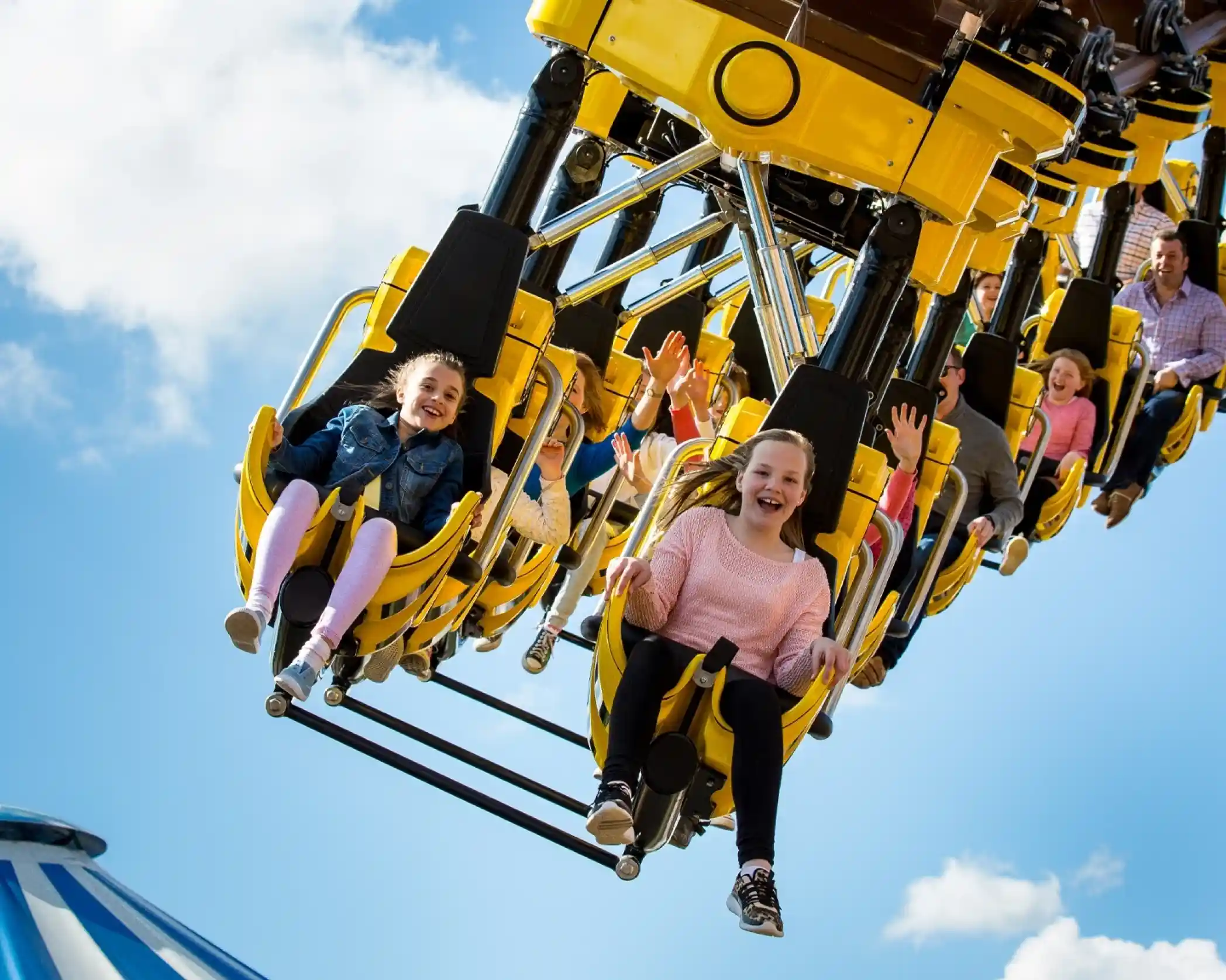 A group of children and adults ride a thrilling amusement park attraction, smiling and raising their hands in excitement against a bright blue sky with scattered clouds.