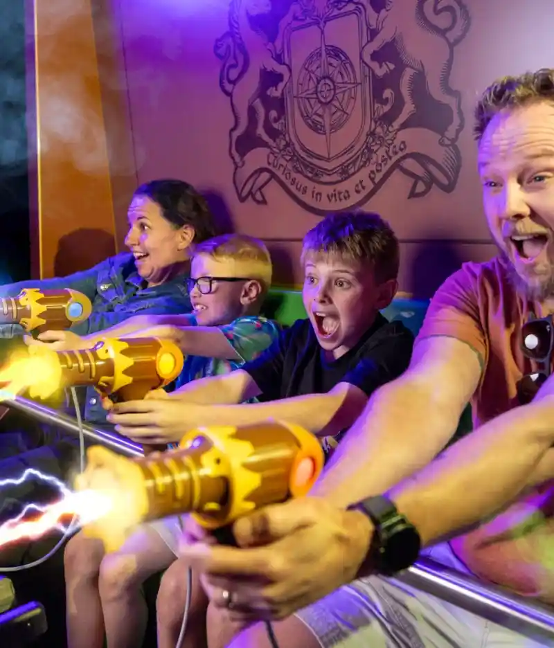 A couple stands on a balcony with a modern home in the background. The man and woman are smiling, dressed casually, with green grass and a landscaped area surrounding the house. The sky is clear and blue.