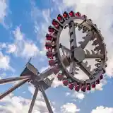 A large amusement park ride featuring a rotating circular structure with seats attached, set against a blue sky with fluffy white clouds.