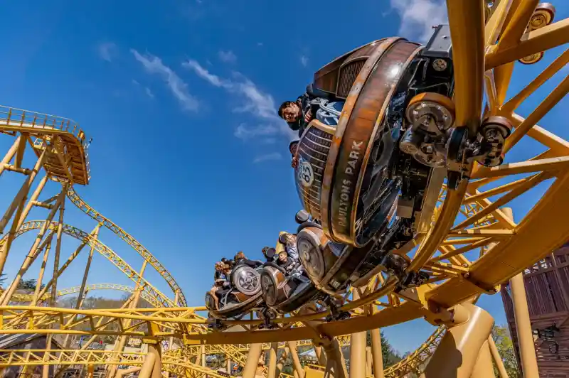 A roller coaster with vintage-style cars zooms through a loop under a clear blue sky. Riders are enjoying the thrill, with some raising their arms in excitement. The track is a bright yellow, contrasting with the surrounding landscape.