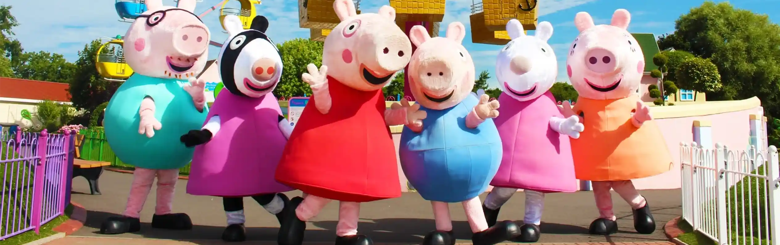 A group of animated characters from a children's show stands together, wearing colorful outfits. They are set against a backdrop of amusement park attractions, including colorful hot air balloons. The characters are smiling and appear cheerful.