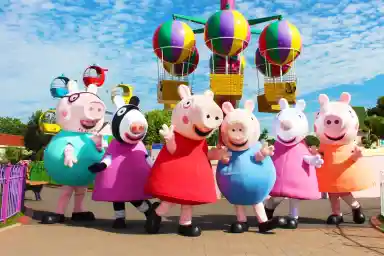 A group of animated characters from a children's show stands together, wearing colorful outfits. They are set against a backdrop of amusement park attractions, including colorful hot air balloons. The characters are smiling and appear cheerful.