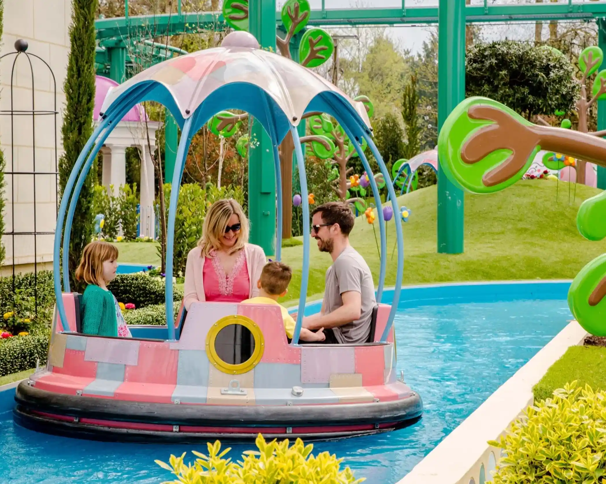 A family enjoys a colorful ride in a circular boat on a water attraction, surrounded by lush greenery and whimsical scenery. The mother and father sit with two children, smiling and interacting as they navigate the cheerful environment.