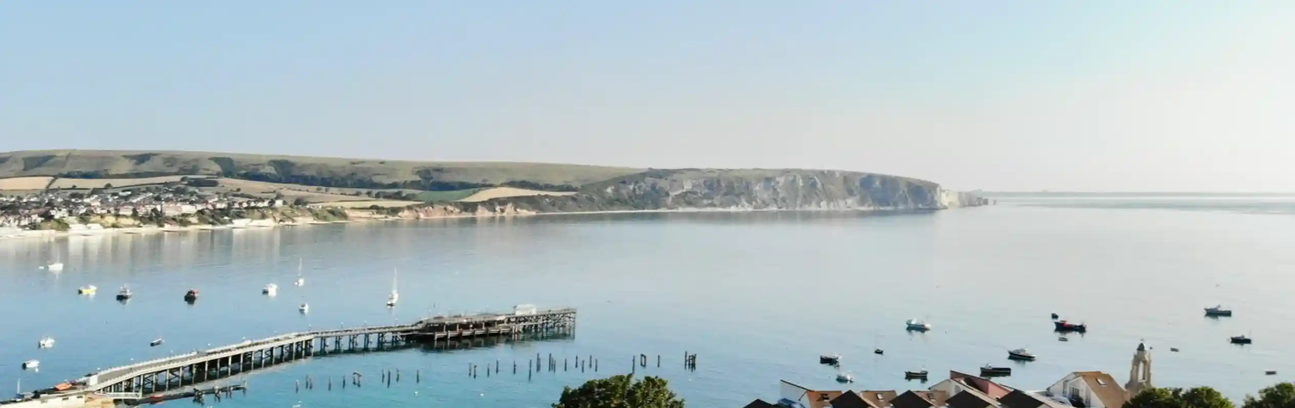 A tranquil coastal scene featuring calm waters with small boats bobbing. In the foreground, lush greenery and a cluster of charming buildings line the shore. A pier extends into the water, while distant hills rise on the horizon under a clear blue sky.