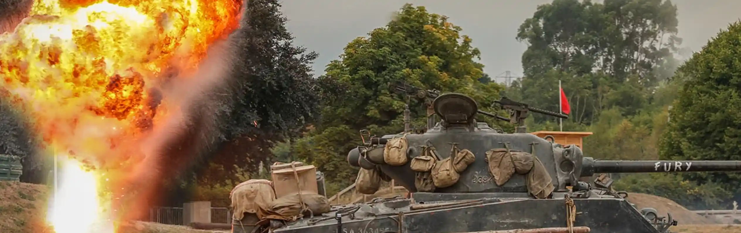 A military tank labeled "FURY" stands on a grassy field, with a large explosion of fire and debris erupting in the background. Thick smoke and orange flames contrast against a cloudy sky and green trees.