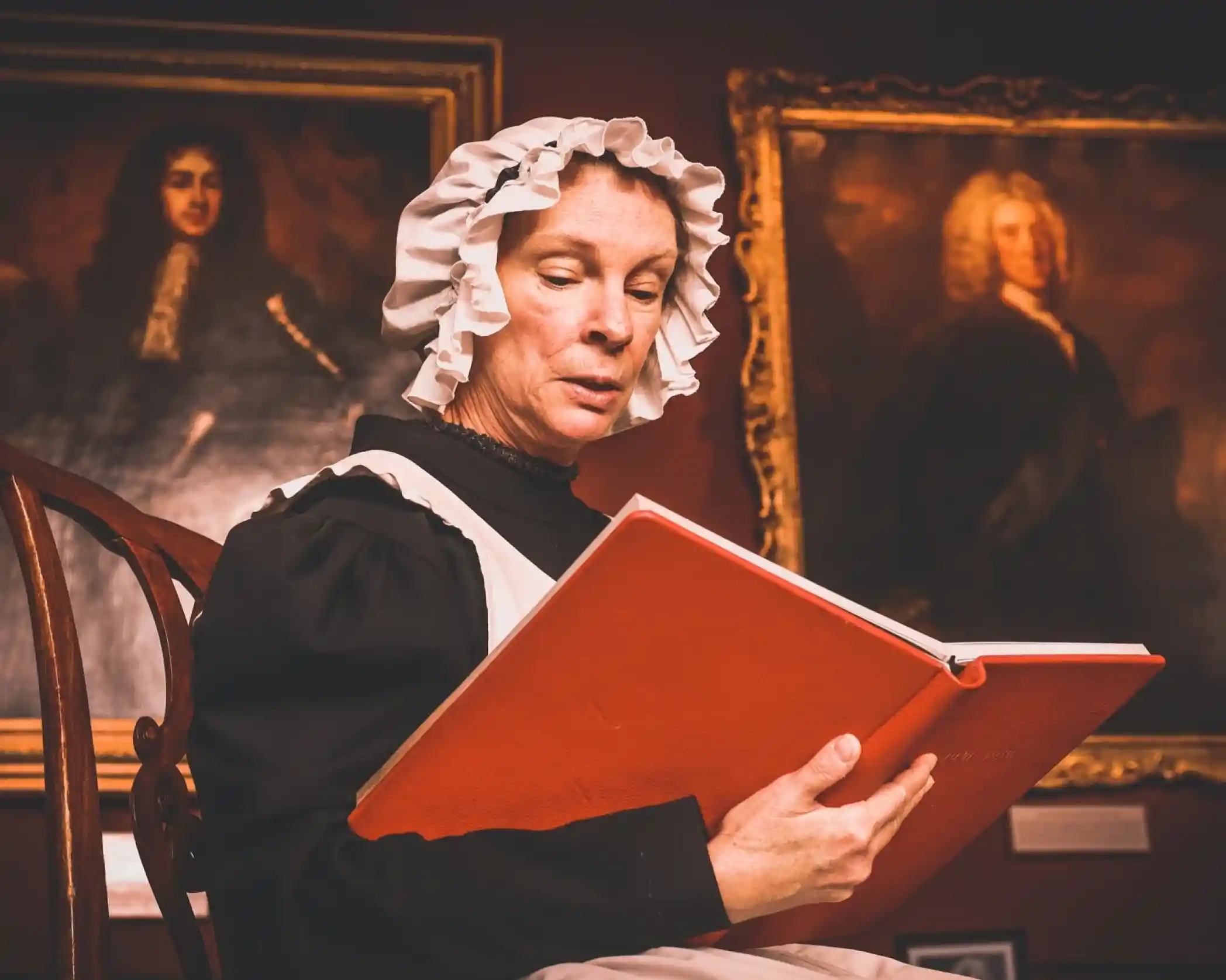 A woman in a period costume, wearing a white cap and black dress, sits in a wooden chair, reading from a large red book. Historical portraits hang on the wall behind her, adding to the setting's vintage atmosphere.