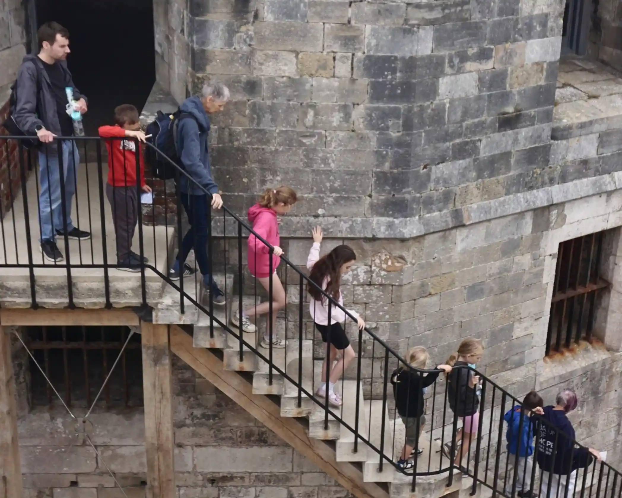 A group of people descending a spiral staircase in a stone tower. They consist of adults and children, some holding drinks and wearing casual clothing. The atmosphere is historic, with visible stone walls and wooden beams.