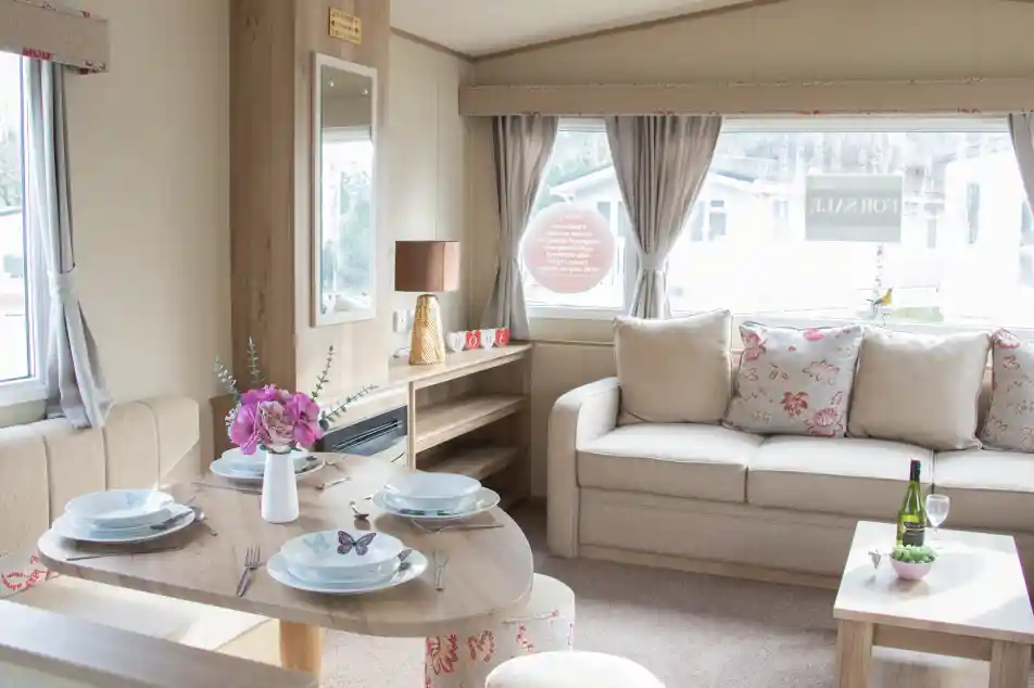 A cozy interior of a holiday home featuring a dining table set for four, a comfortable sofa with floral cushions, and a decorative side table with a wine bottle and glasses. Natural light filters through large windows adorned with curtains.