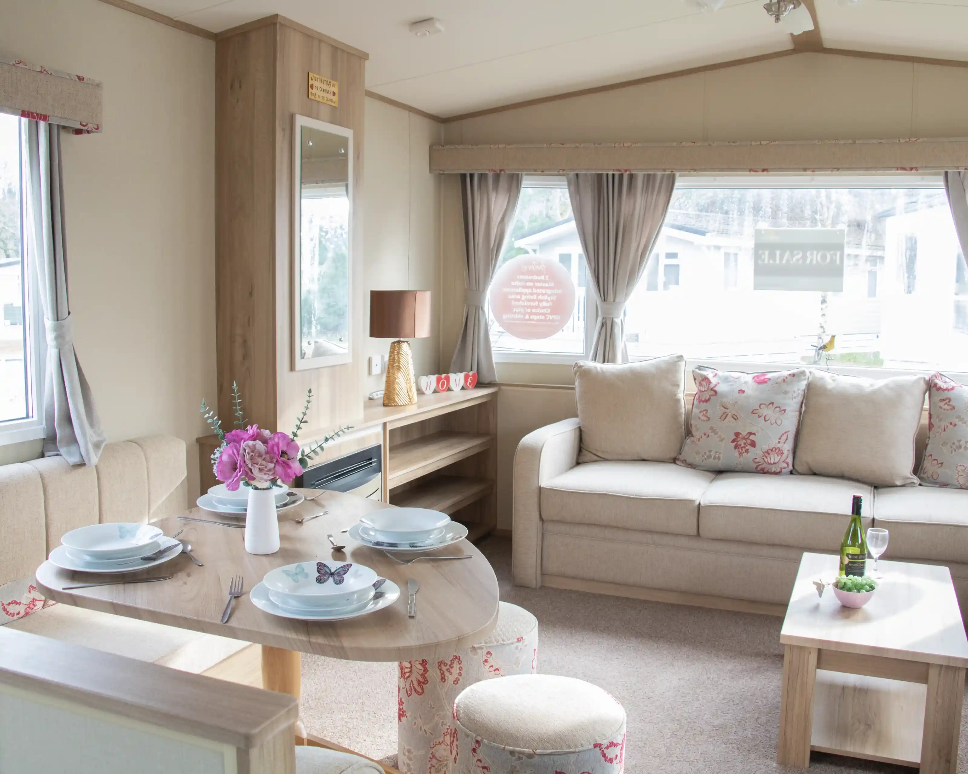 A cozy interior of a holiday home featuring a dining table set for four, a comfortable sofa with floral cushions, and a decorative side table with a wine bottle and glasses. Natural light filters through large windows adorned with curtains.
