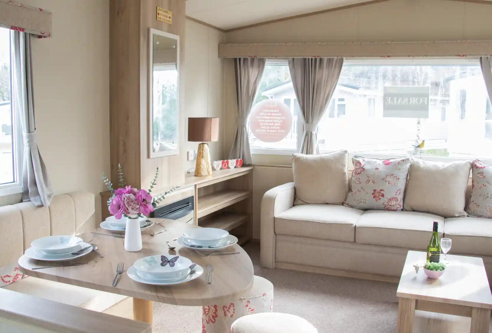 A cozy interior of a holiday home featuring a dining table set for four, a comfortable sofa with floral cushions, and a decorative side table with a wine bottle and glasses. Natural light filters through large windows adorned with curtains.