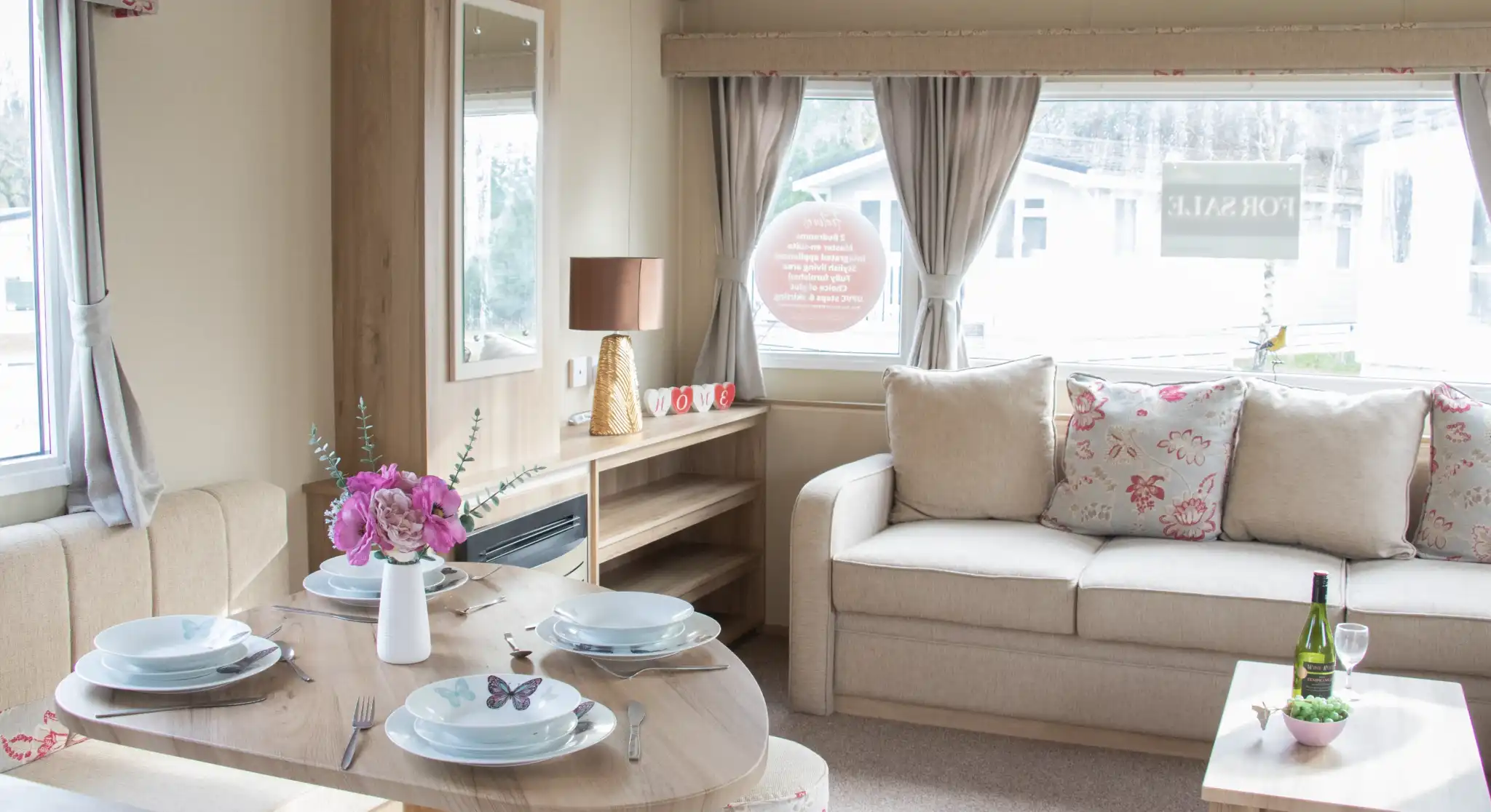 A cozy interior of a holiday home featuring a dining table set for four, a comfortable sofa with floral cushions, and a decorative side table with a wine bottle and glasses. Natural light filters through large windows adorned with curtains.