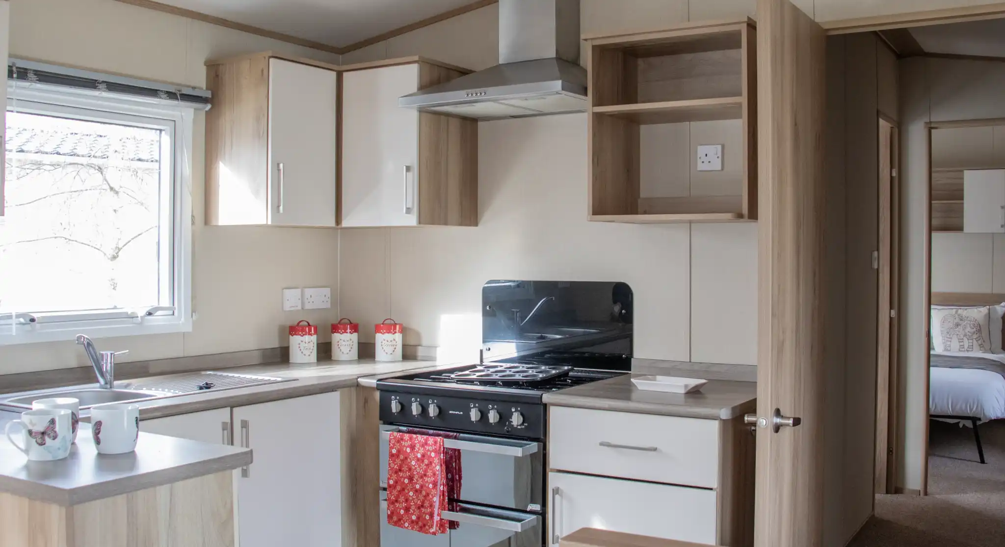 A modern kitchen with wooden cabinetry, a stove and oven, a sink, and countertop space. Light streams in through a window, and there are decorative items on the countertop. In the background, a bedroom area is visible through an open door.