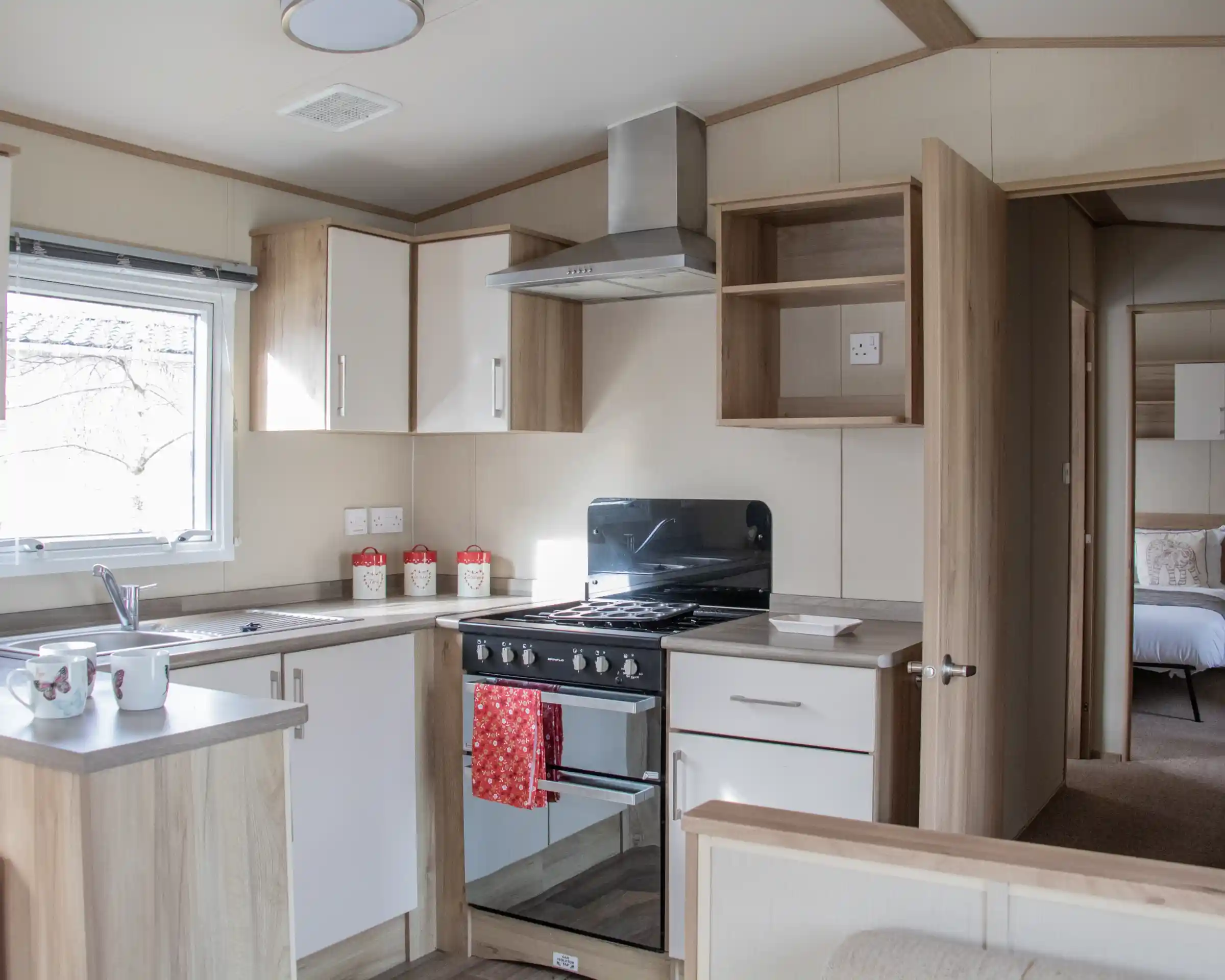 A modern kitchen with wooden cabinetry, a stove and oven, a sink, and countertop space. Light streams in through a window, and there are decorative items on the countertop. In the background, a bedroom area is visible through an open door.