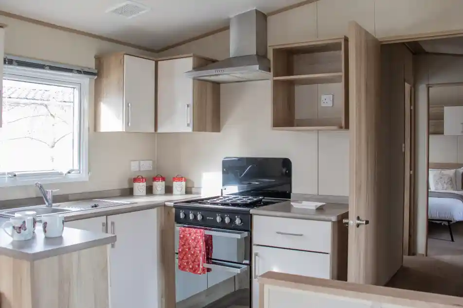 A modern kitchen with wooden cabinetry, a stove and oven, a sink, and countertop space. Light streams in through a window, and there are decorative items on the countertop. In the background, a bedroom area is visible through an open door.