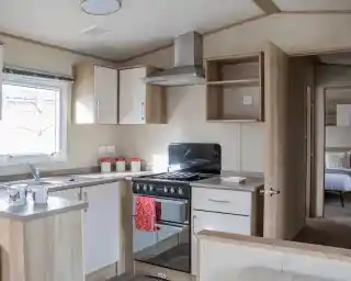 A modern kitchen with wooden cabinetry, a stove and oven, a sink, and countertop space. Light streams in through a window, and there are decorative items on the countertop. In the background, a bedroom area is visible through an open door.