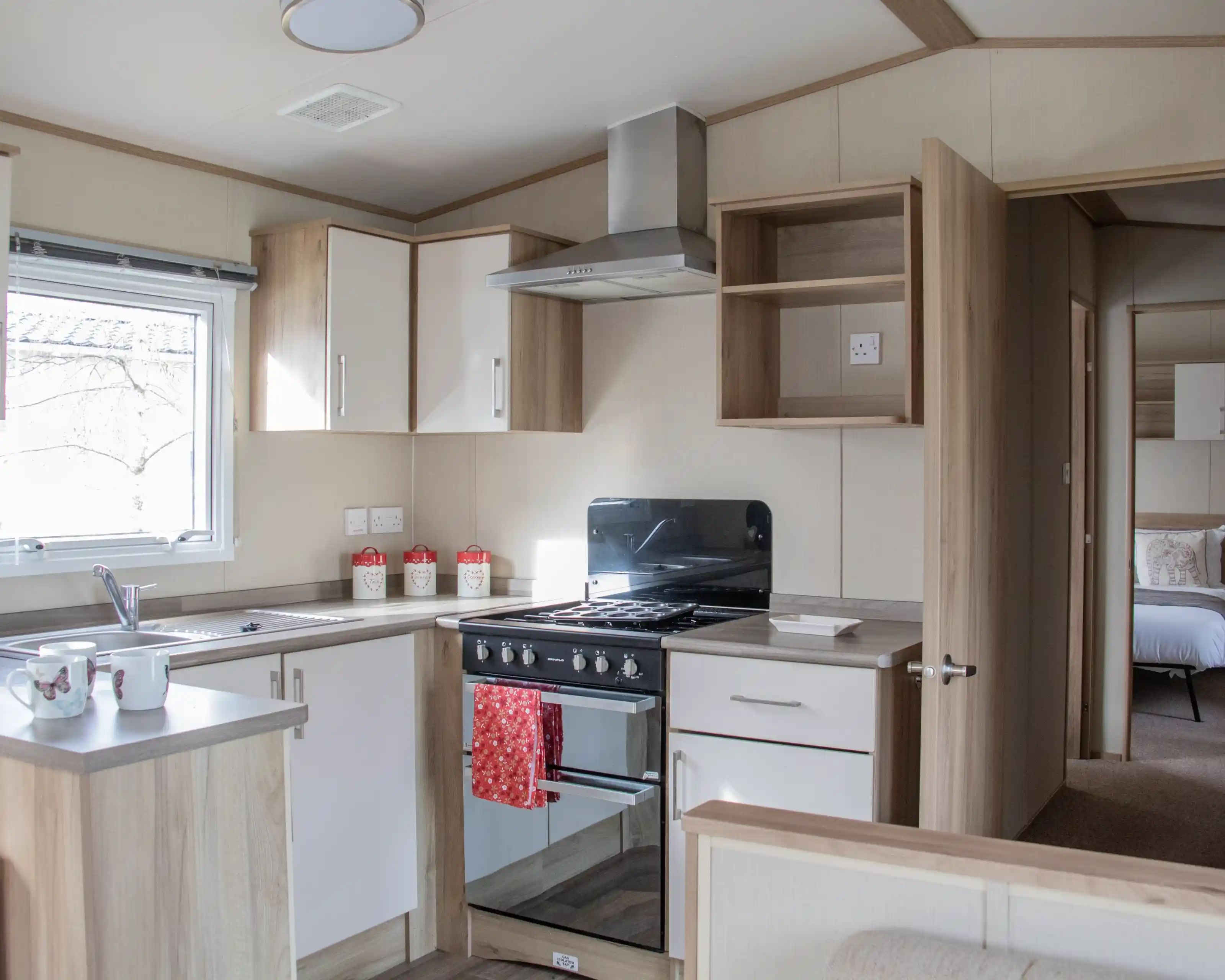 A modern kitchen with wooden cabinetry, a stove and oven, a sink, and countertop space. Light streams in through a window, and there are decorative items on the countertop. In the background, a bedroom area is visible through an open door.