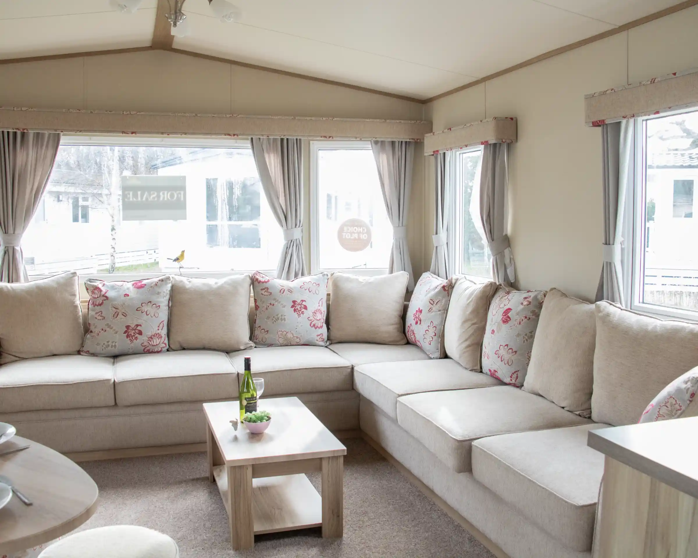 A cozy living area featuring a large L-shaped sofa with floral-patterned cushions. There is a small wooden coffee table in front, holding a bottle of wine and a snack plate. Large windows let in natural light, revealing a view of the outdoors.