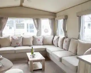 A cozy living area featuring a large L-shaped sofa with floral-patterned cushions. There is a small wooden coffee table in front, holding a bottle of wine and a snack plate. Large windows let in natural light, revealing a view of the outdoors.