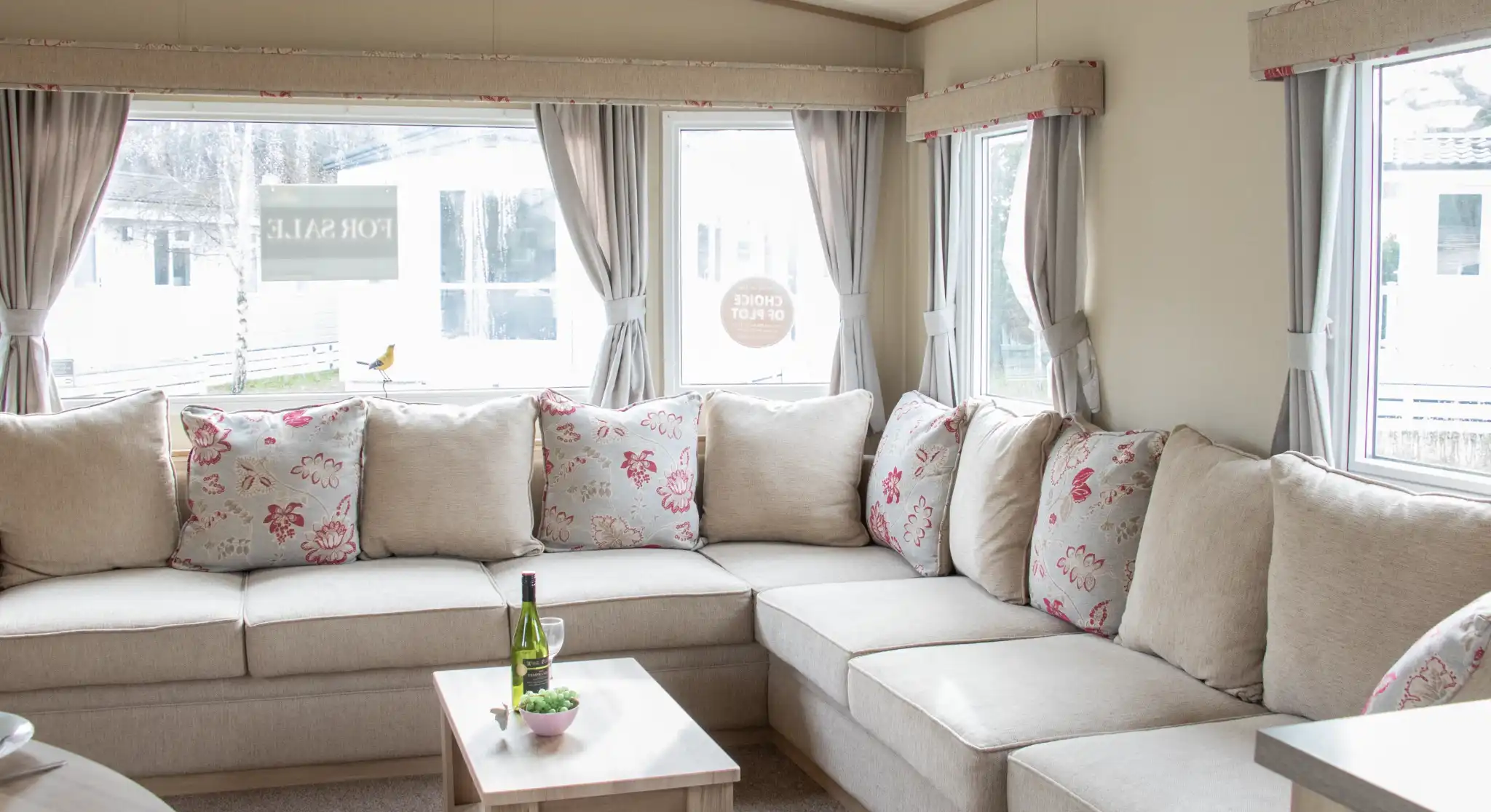 A cozy living area featuring a large L-shaped sofa with floral-patterned cushions. There is a small wooden coffee table in front, holding a bottle of wine and a snack plate. Large windows let in natural light, revealing a view of the outdoors.