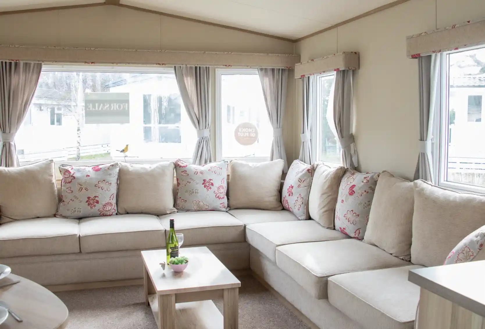 A cozy living area featuring a large L-shaped sofa with floral-patterned cushions. There is a small wooden coffee table in front, holding a bottle of wine and a snack plate. Large windows let in natural light, revealing a view of the outdoors.