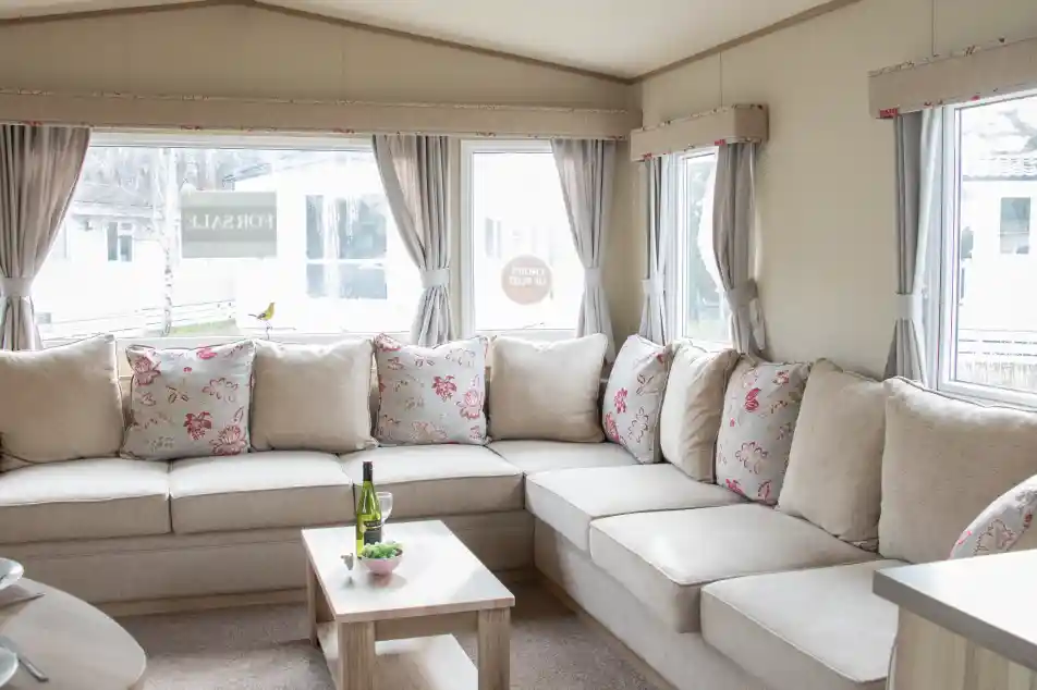 A cozy living area featuring a large L-shaped sofa with floral-patterned cushions. There is a small wooden coffee table in front, holding a bottle of wine and a snack plate. Large windows let in natural light, revealing a view of the outdoors.