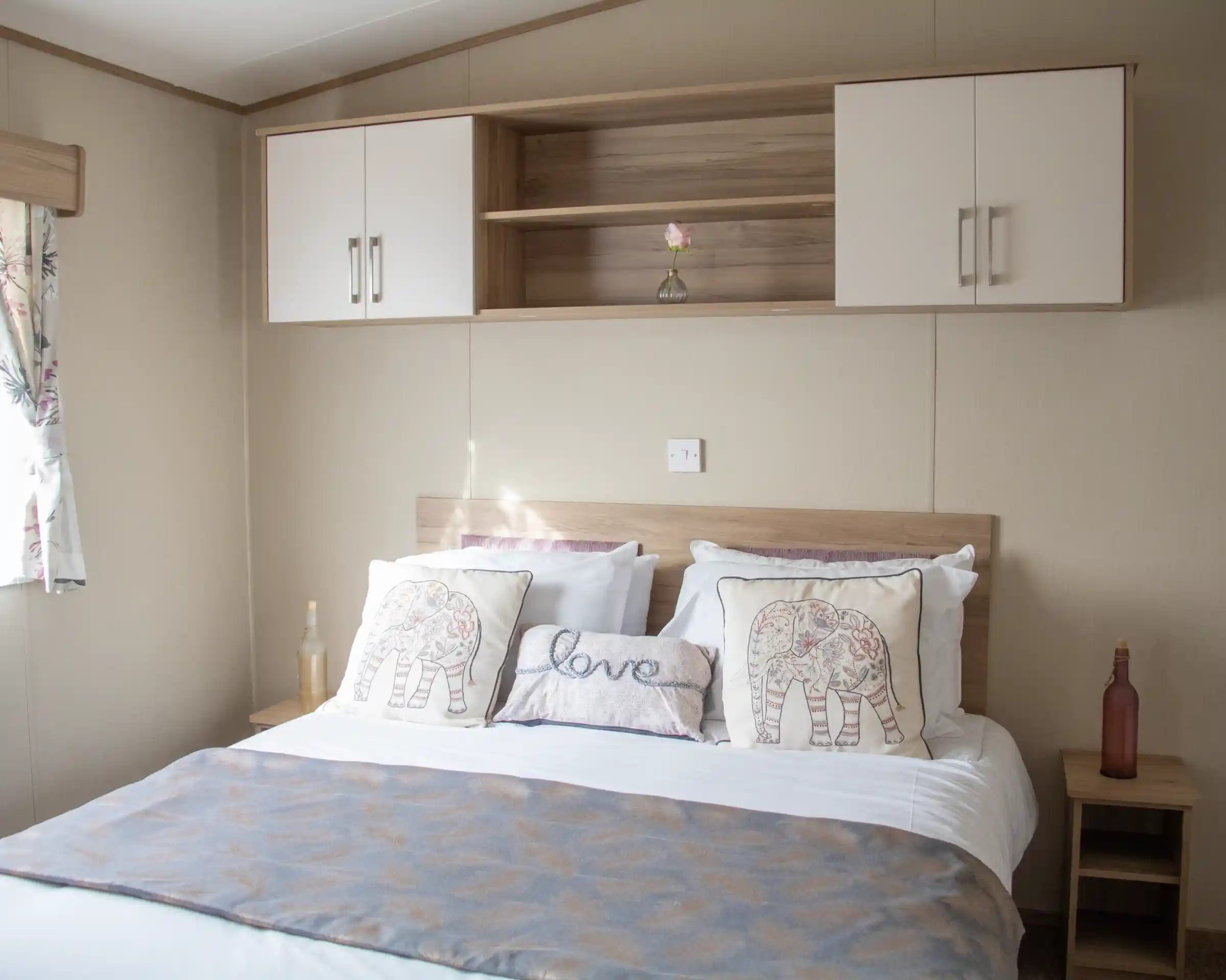 A neatly made bed with white linens and decorative pillows featuring elephant designs. A light gray throw blanket is draped across the bed. A wooden shelf above the bed holds a single flower in a vase and two closed cabinets. Natural light filters in from a nearby window with floral curtains.