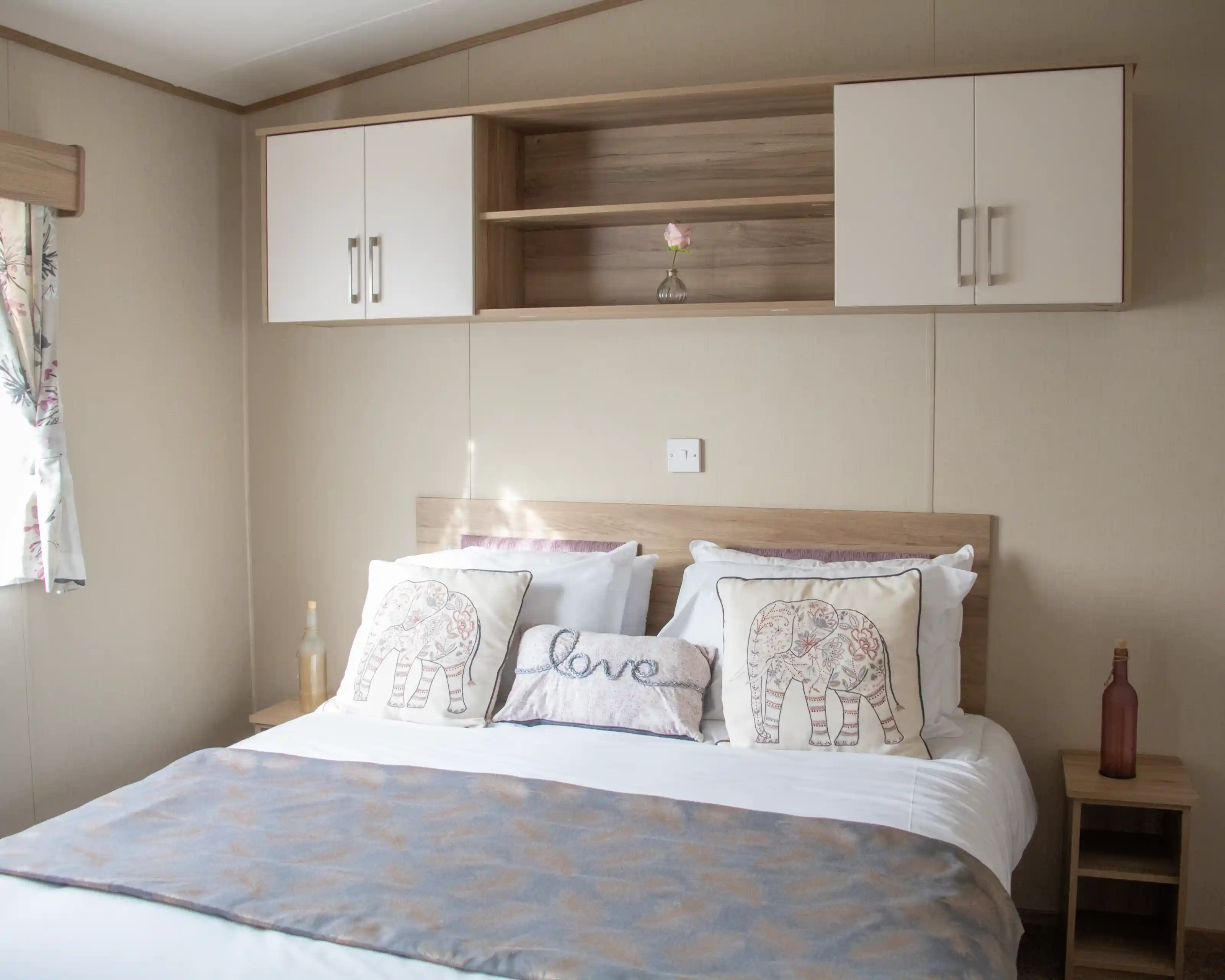 A neatly made bed with white linens and decorative pillows featuring elephant designs. A light gray throw blanket is draped across the bed. A wooden shelf above the bed holds a single flower in a vase and two closed cabinets. Natural light filters in from a nearby window with floral curtains.