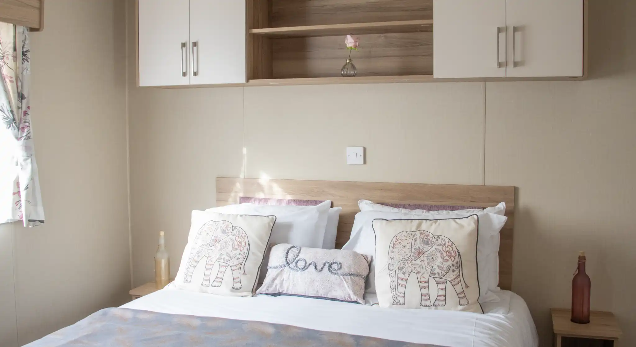 A neatly made bed with white linens and decorative pillows featuring elephant designs. A light gray throw blanket is draped across the bed. A wooden shelf above the bed holds a single flower in a vase and two closed cabinets. Natural light filters in from a nearby window with floral curtains.