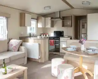 A cozy interior of a modern holiday home featuring a comfortable sofa with floral cushions, a round dining table set with plates, and a kitchenette with a stove and cabinets. Natural light floods the space, enhancing the warm wood tones and soft decor.