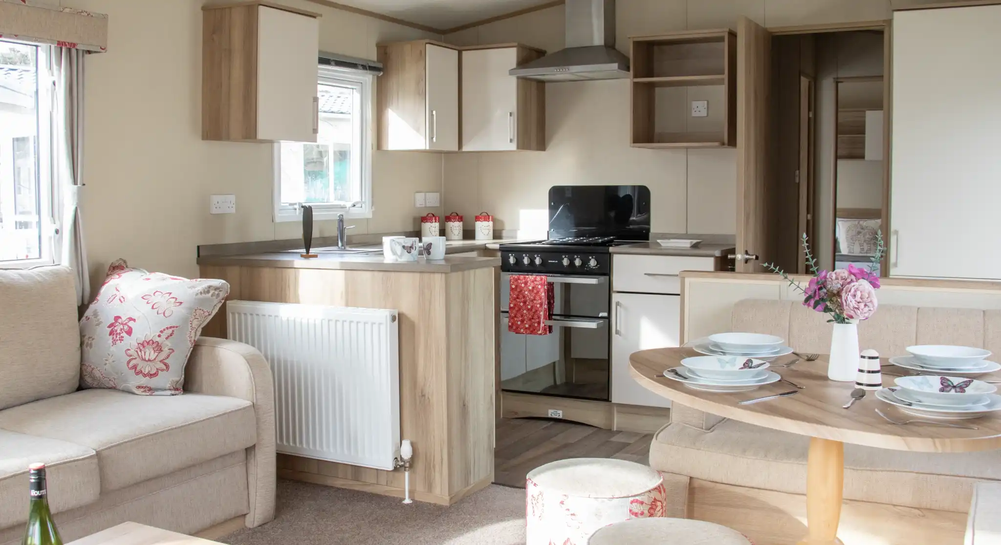 A cozy interior of a modern holiday home featuring a comfortable sofa with floral cushions, a round dining table set with plates, and a kitchenette with a stove and cabinets. Natural light floods the space, enhancing the warm wood tones and soft decor.
