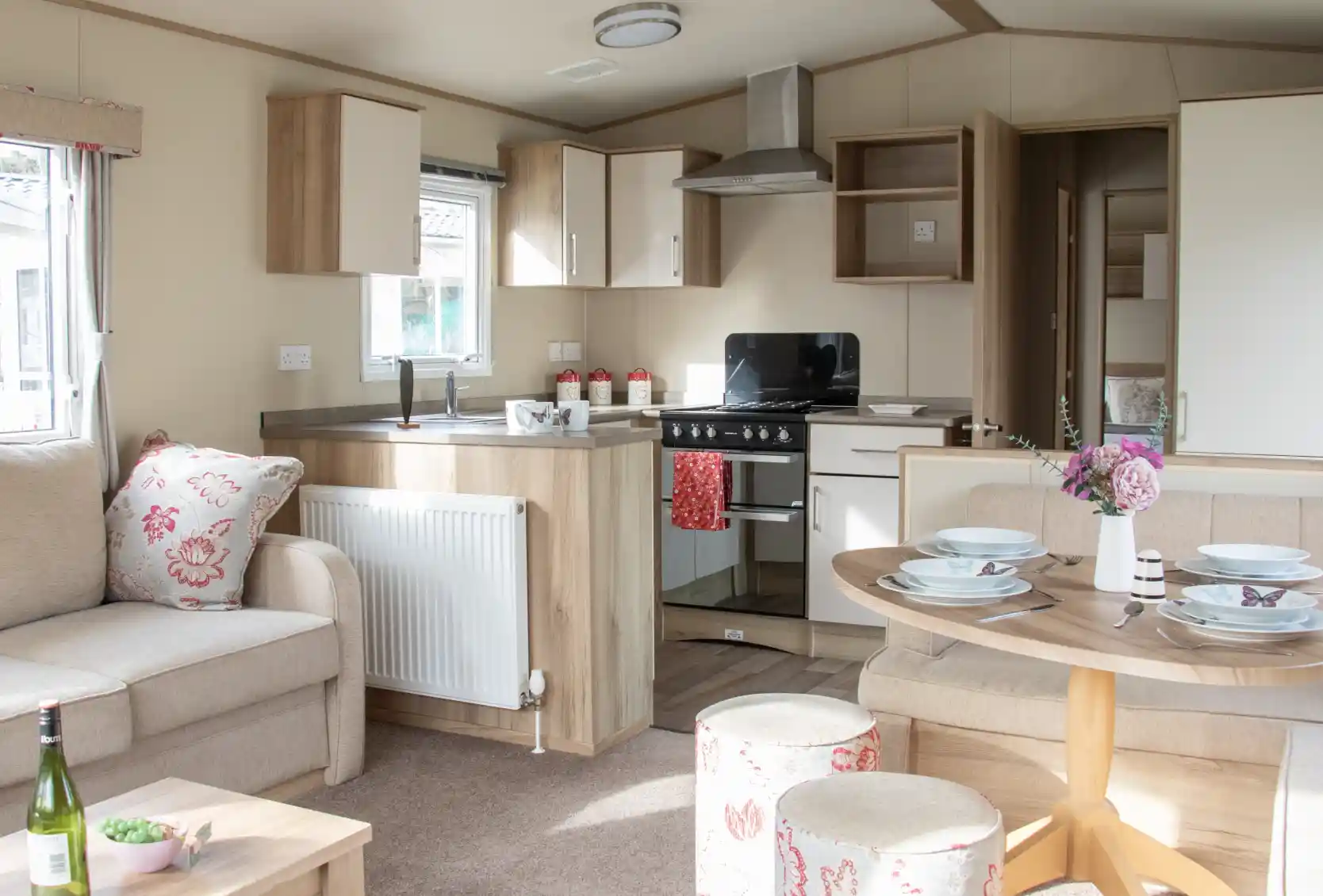 A cozy interior of a modern holiday home featuring a comfortable sofa with floral cushions, a round dining table set with plates, and a kitchenette with a stove and cabinets. Natural light floods the space, enhancing the warm wood tones and soft decor.