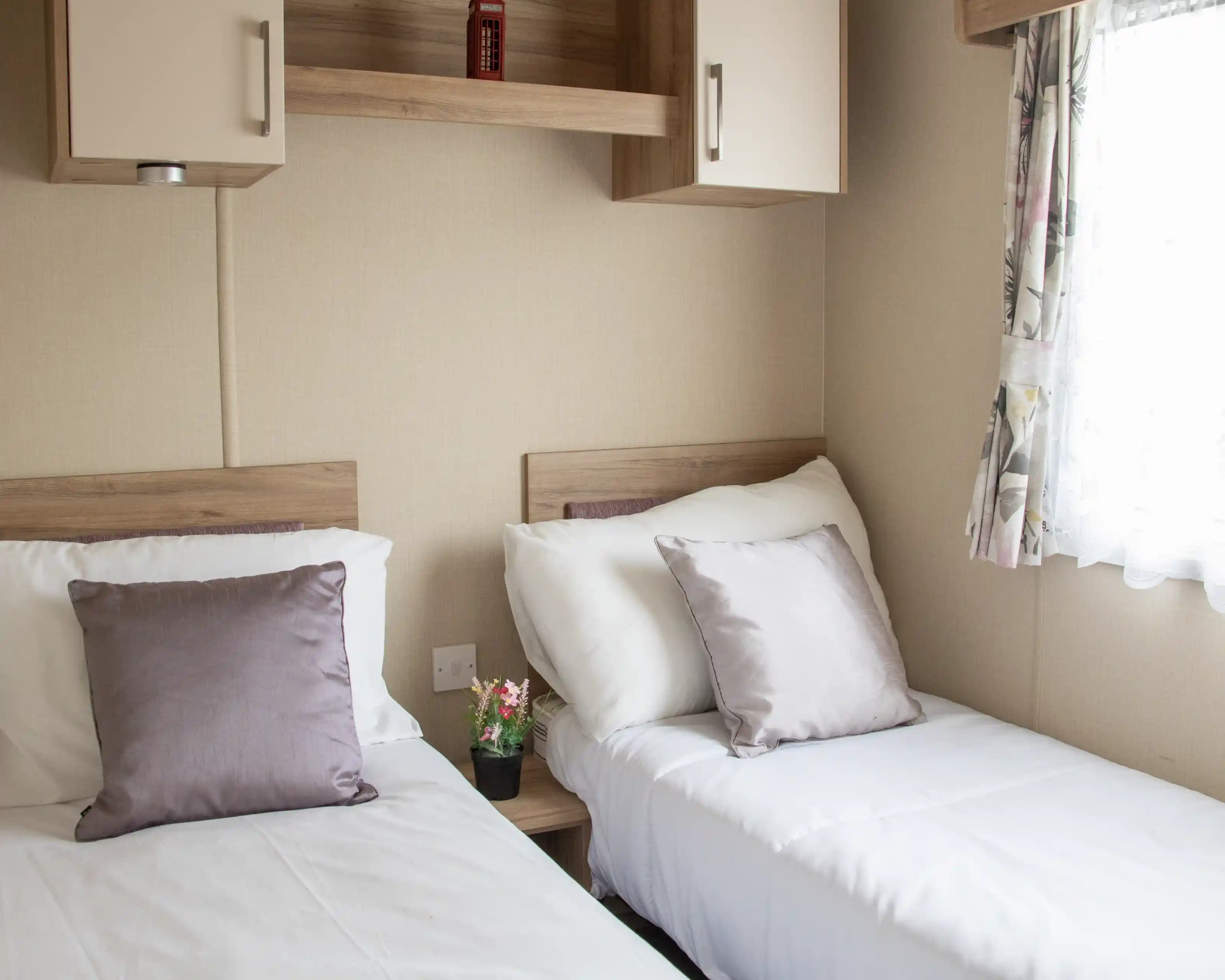 Two twin beds with white linens and soft purple pillows are positioned in a cozy room. A small potted plant sits on a bedside table, and a window with floral curtains allows natural light to enter. Shelves above the beds add to the room's functional decor.