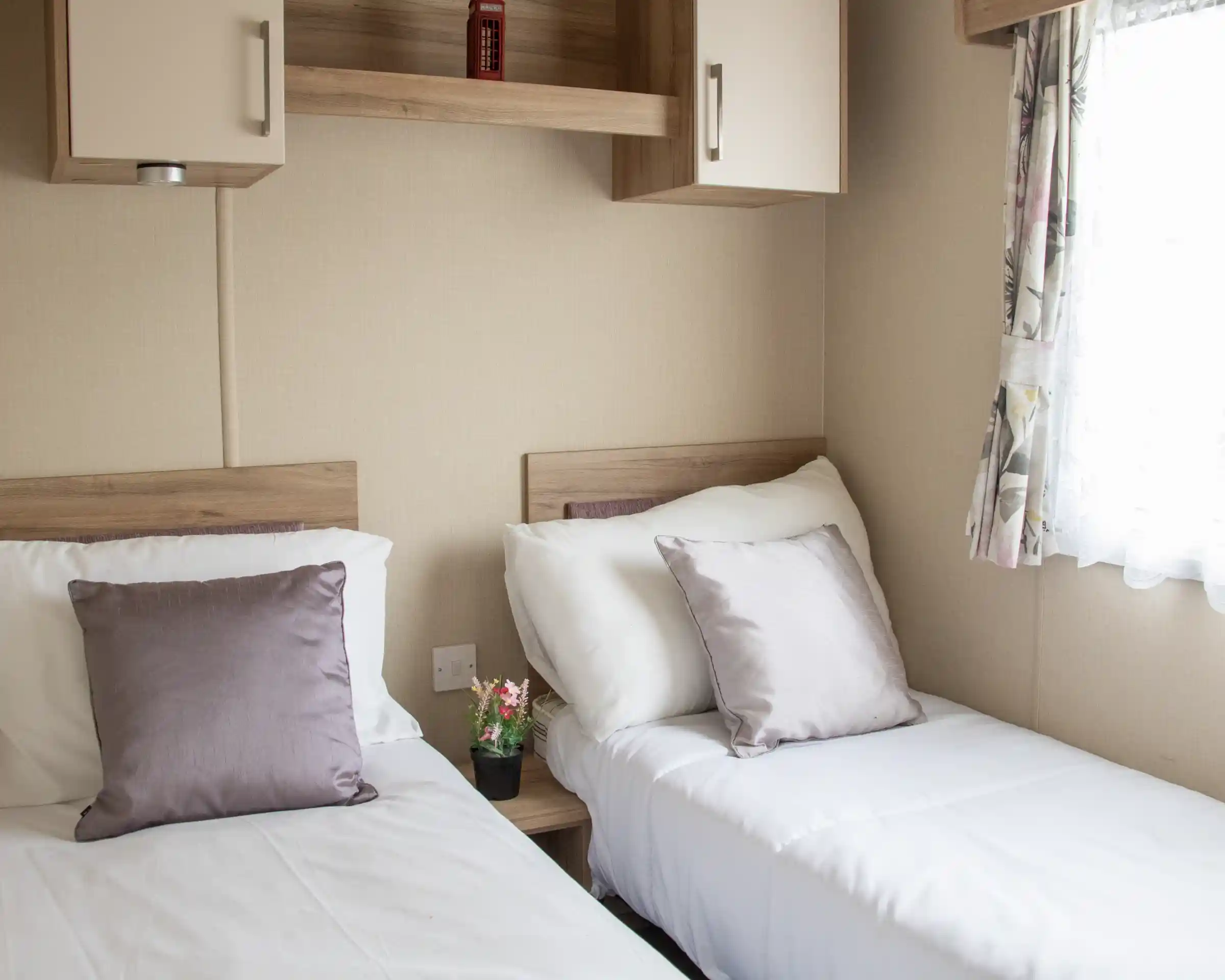 Two twin beds with white linens and soft purple pillows are positioned in a cozy room. A small potted plant sits on a bedside table, and a window with floral curtains allows natural light to enter. Shelves above the beds add to the room's functional decor.