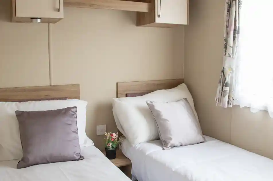 Two twin beds with white linens and soft purple pillows are positioned in a cozy room. A small potted plant sits on a bedside table, and a window with floral curtains allows natural light to enter. Shelves above the beds add to the room's functional decor.