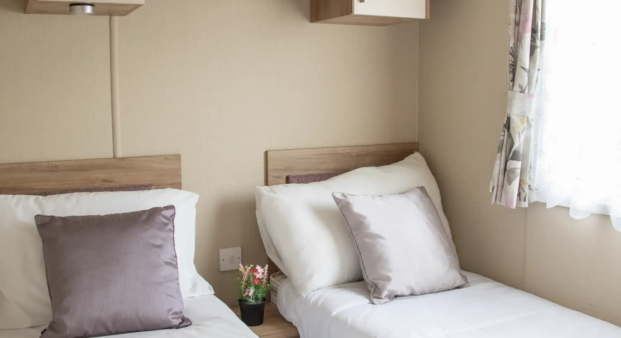 Two twin beds with white linens and soft purple pillows are positioned in a cozy room. A small potted plant sits on a bedside table, and a window with floral curtains allows natural light to enter. Shelves above the beds add to the room's functional decor.