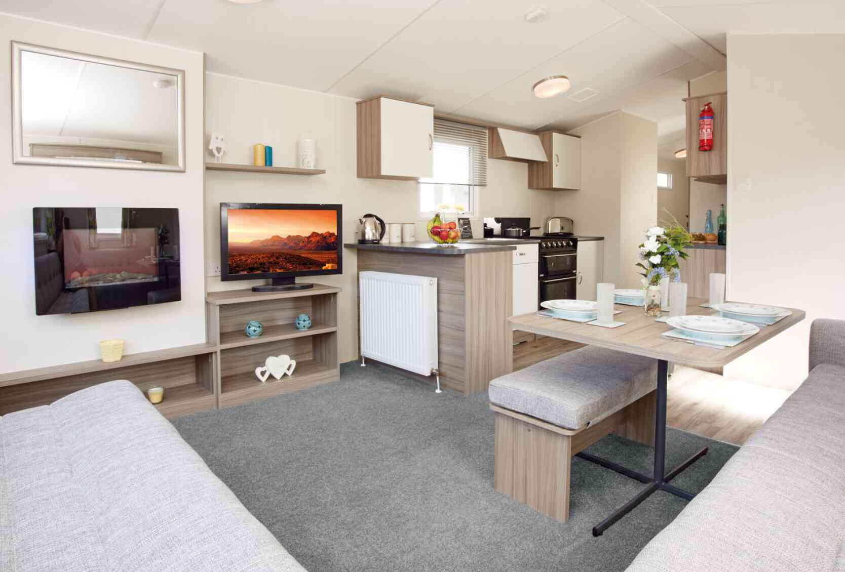 The image shows the interior of a bright, modern caravan or mobile home, featuring an open-plan lounge, dining, and kitchen area. In the lounge section, there’s a wall-mounted electric fireplace with a reflective glass surface, a flat-screen TV displaying a vivid desert landscape, and light wood shelving holding decorative items, including teal candle holders and a white heart-shaped ornament. The dining area has a wooden table set with four plates, glasses, placemats, and a vase of fresh flowers, accompanied by cushioned benches upholstered in light grey fabric. The kitchen, located behind the dining space, has light wood and white cabinetry, a countertop with a kettle and a bowl of fruit, and a black freestanding oven with a gas hob. A fire extinguisher is mounted on the wall in the background. The space is decorated in neutral tones with light grey carpet in the lounge and wood-effect flooring in the kitchen and dining areas.