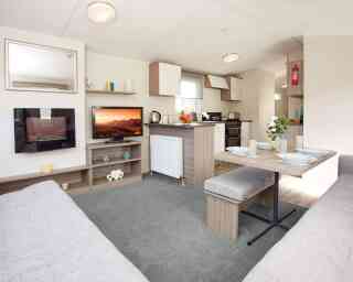 The image shows the interior of a bright, modern caravan or mobile home, featuring an open-plan lounge, dining, and kitchen area. In the lounge section, there’s a wall-mounted electric fireplace with a reflective glass surface, a flat-screen TV displaying a vivid desert landscape, and light wood shelving holding decorative items, including teal candle holders and a white heart-shaped ornament. The dining area has a wooden table set with four plates, glasses, placemats, and a vase of fresh flowers, accompanied by cushioned benches upholstered in light grey fabric. The kitchen, located behind the dining space, has light wood and white cabinetry, a countertop with a kettle and a bowl of fruit, and a black freestanding oven with a gas hob. A fire extinguisher is mounted on the wall in the background. The space is decorated in neutral tones with light grey carpet in the lounge and wood-effect flooring in the kitchen and dining areas.