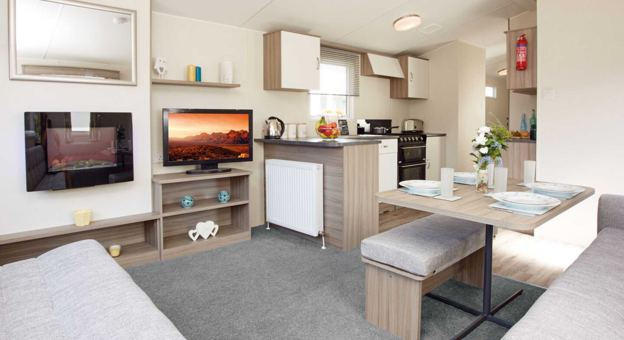 The image shows the interior of a bright, modern caravan or mobile home, featuring an open-plan lounge, dining, and kitchen area. In the lounge section, there’s a wall-mounted electric fireplace with a reflective glass surface, a flat-screen TV displaying a vivid desert landscape, and light wood shelving holding decorative items, including teal candle holders and a white heart-shaped ornament. The dining area has a wooden table set with four plates, glasses, placemats, and a vase of fresh flowers, accompanied by cushioned benches upholstered in light grey fabric. The kitchen, located behind the dining space, has light wood and white cabinetry, a countertop with a kettle and a bowl of fruit, and a black freestanding oven with a gas hob. A fire extinguisher is mounted on the wall in the background. The space is decorated in neutral tones with light grey carpet in the lounge and wood-effect flooring in the kitchen and dining areas.